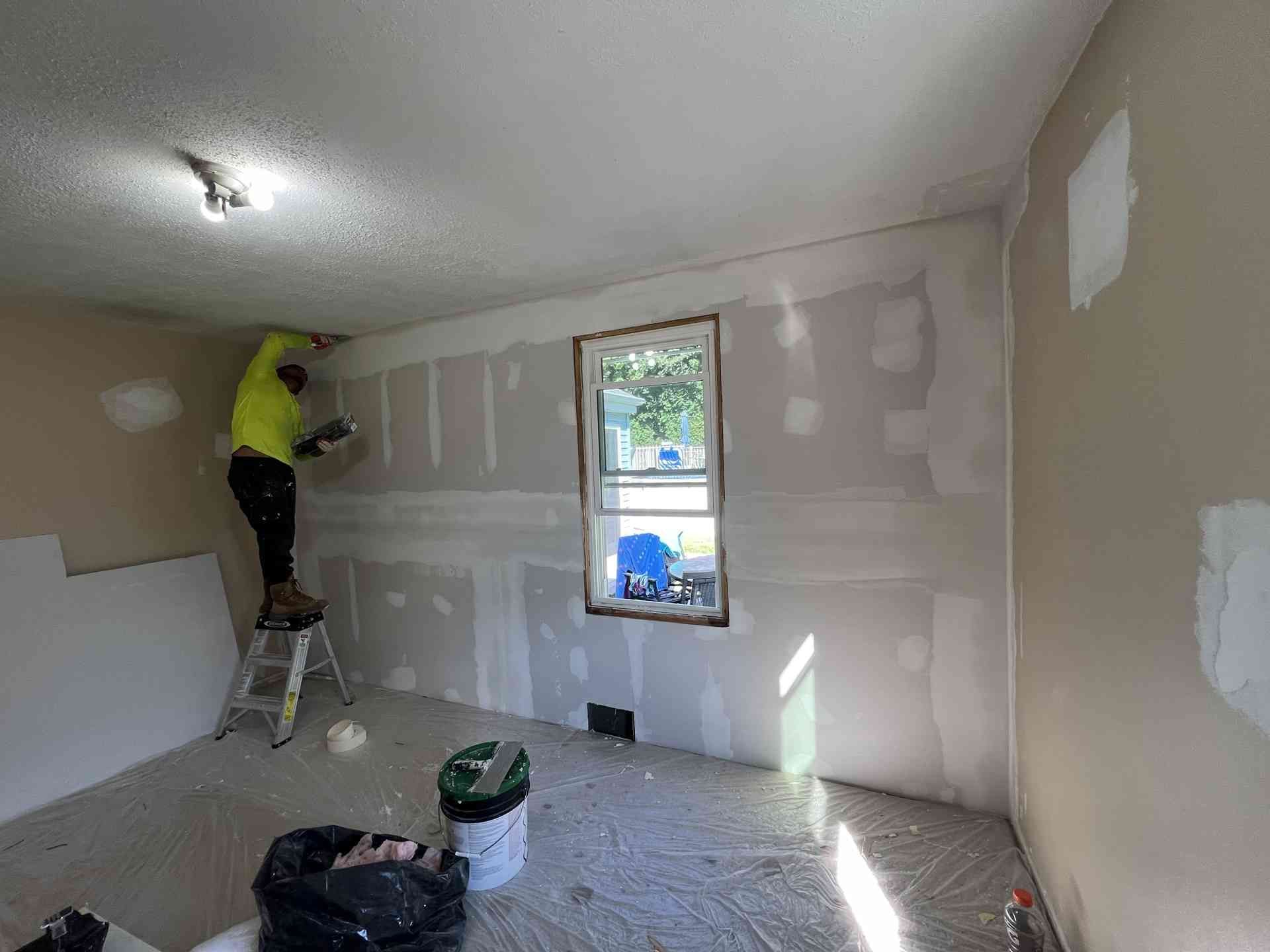 Person painting drywall in a small room with a window and step ladder.