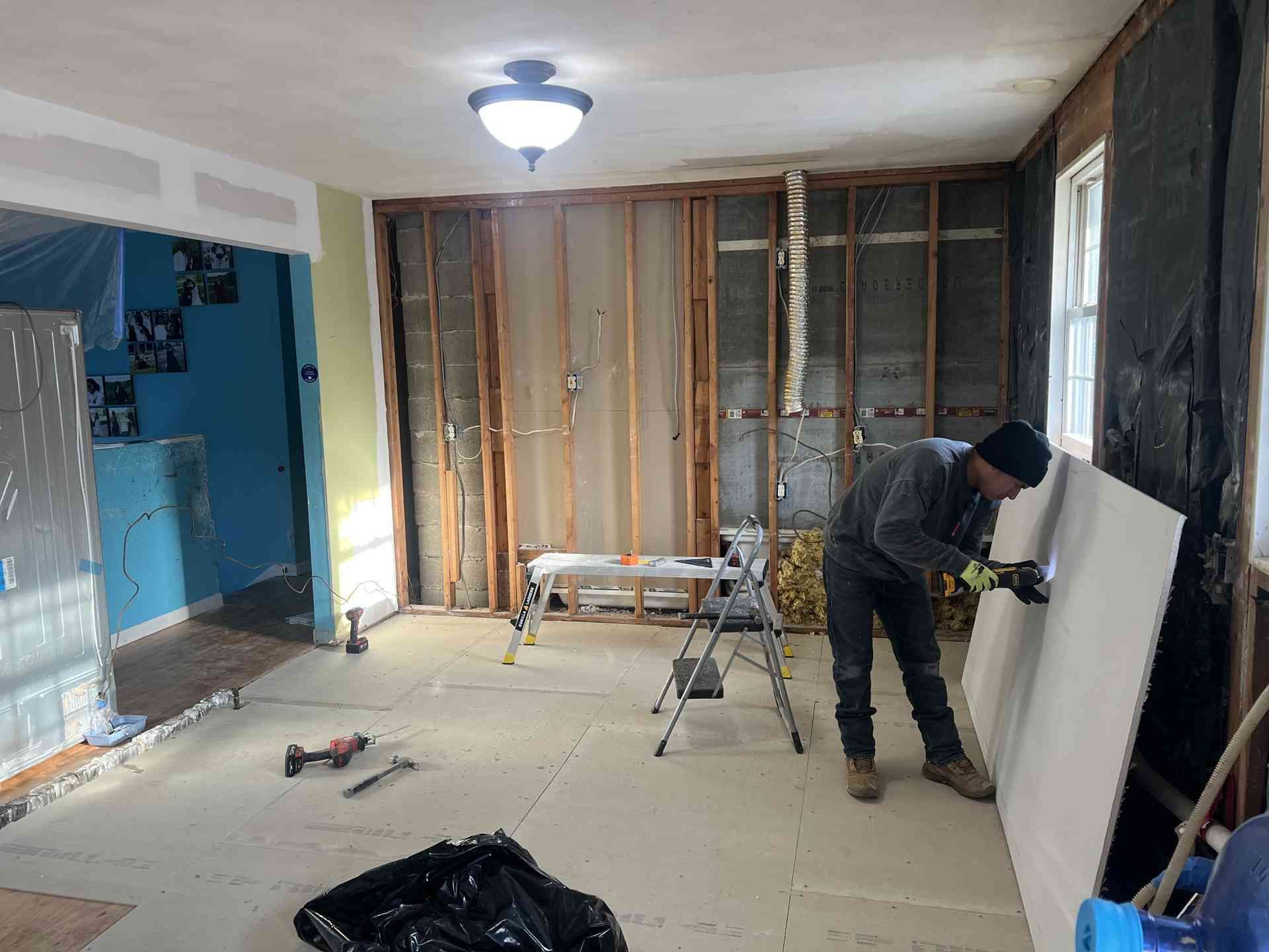 Person installing drywall in an unfinished room with exposed studs and blue-painted adjacent space.