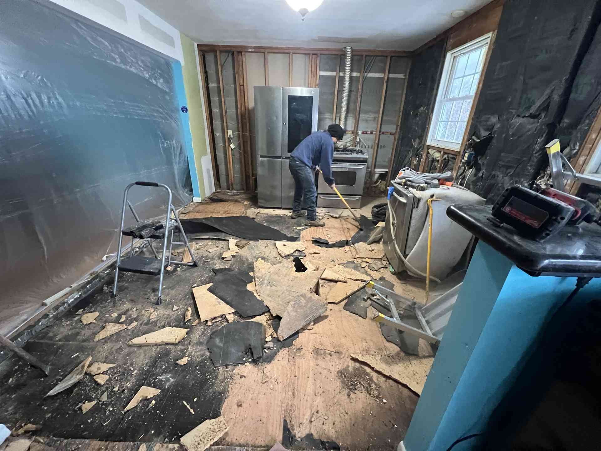 Person cleaning debris from a damaged room with broken floor tiles and plastic-covered walls