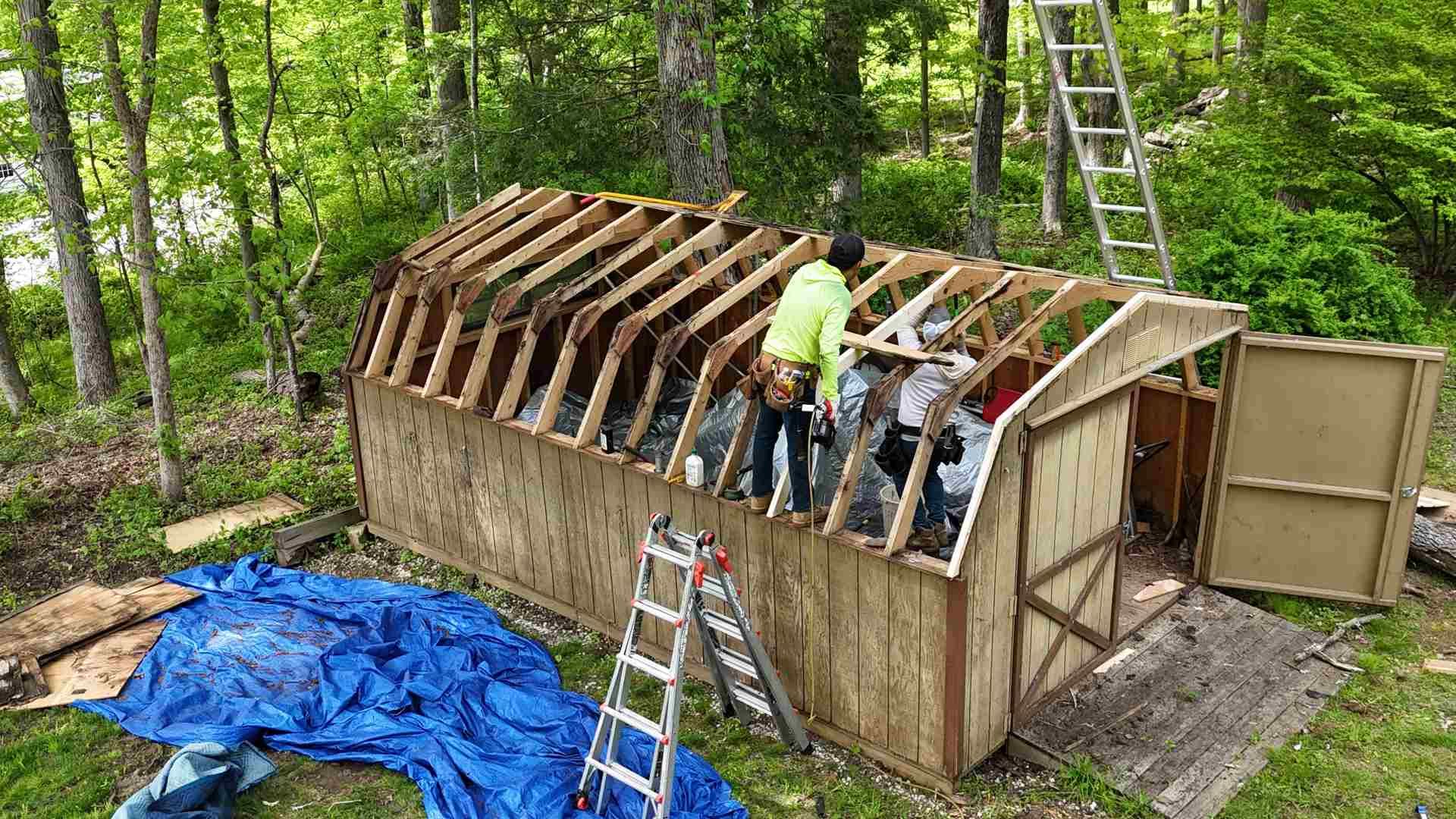 Two people building a wooden shed roof in a wooded area, with ladders and tarps nearby.