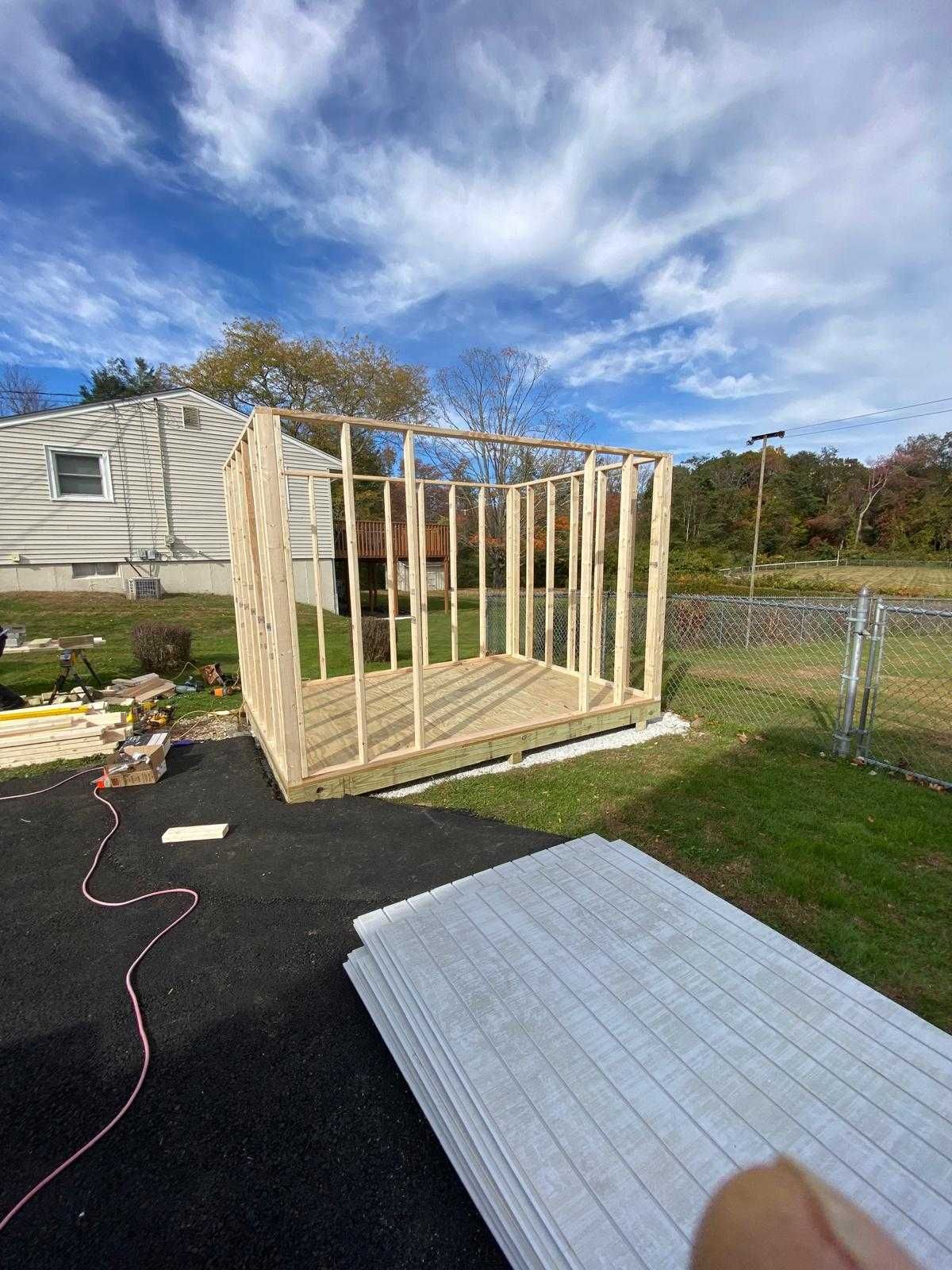 Framed wooden shed under construction beside a house, with lumber, black tarp, and a white panel on the grass.