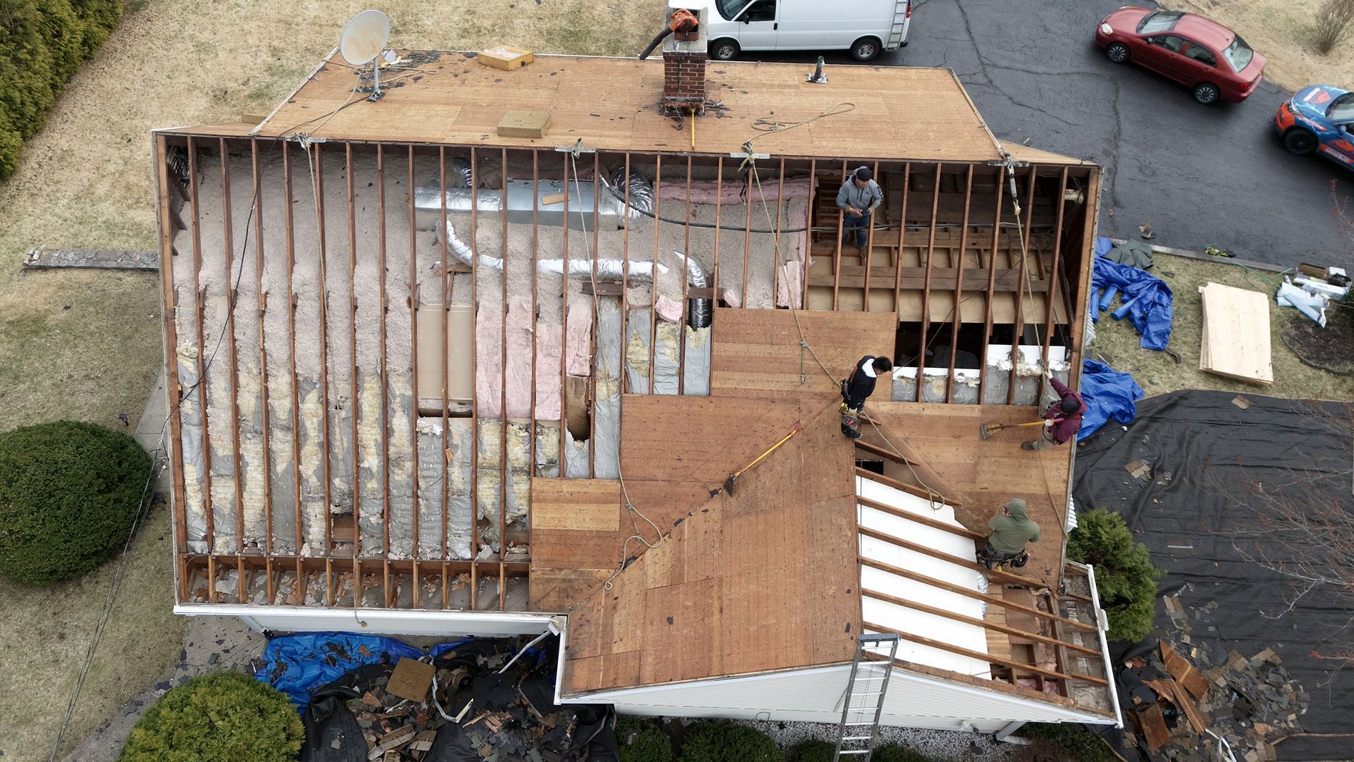 Aerial view of a house with a partially stripped roof, exposing rafters and plywood during repairs