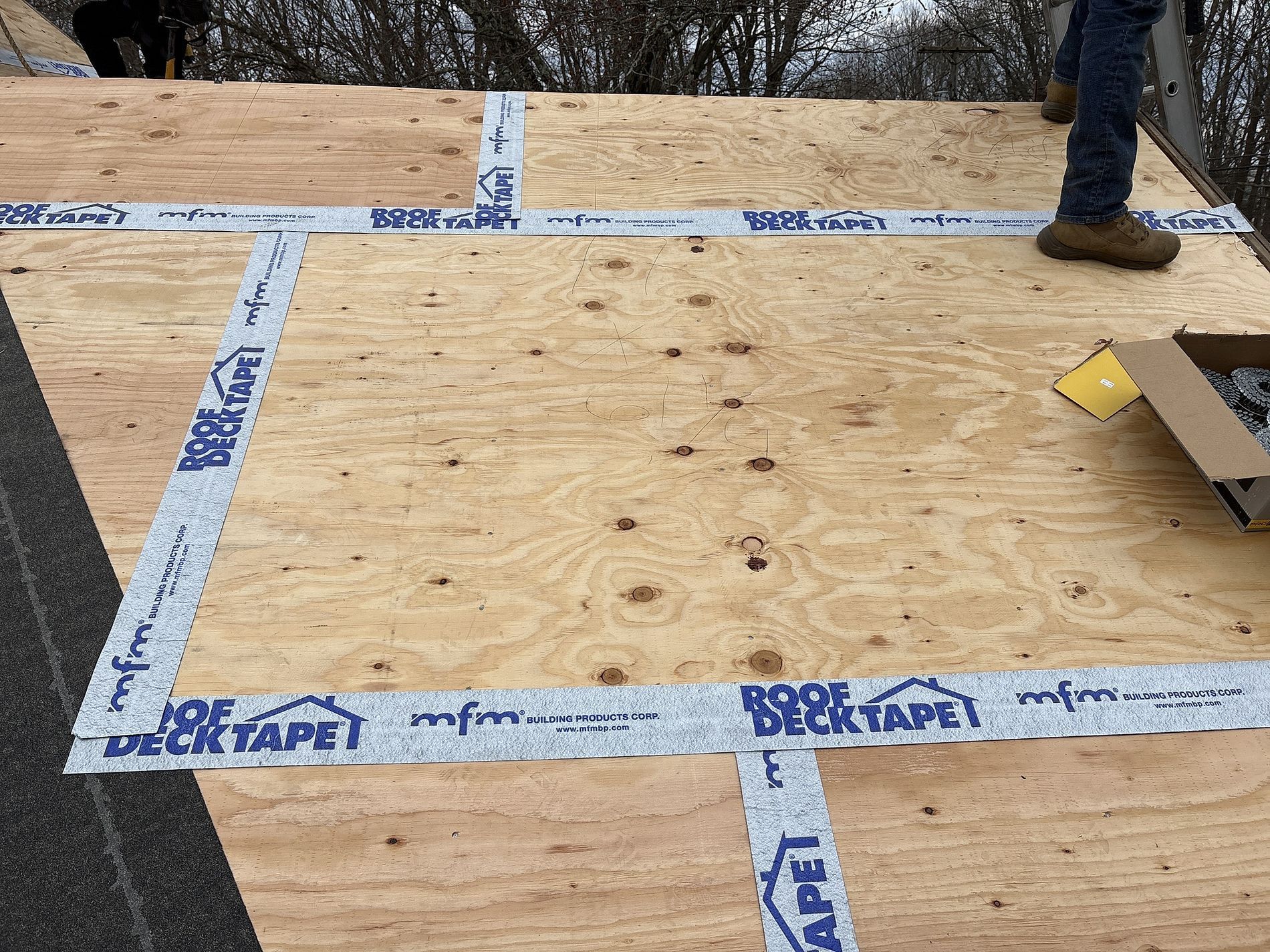 Plywood roof deck with blue tape seams and a worker’s boot at the edge of the frame
