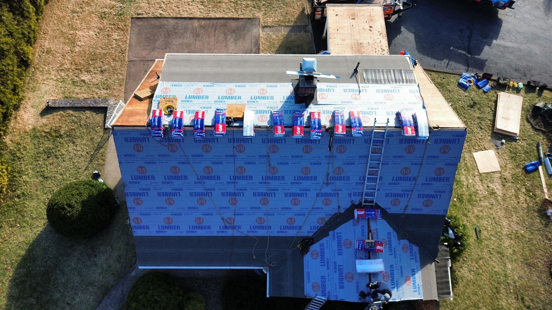 Aerial view of a house roof under construction with blue roofing underlayment and workers near the ridge.