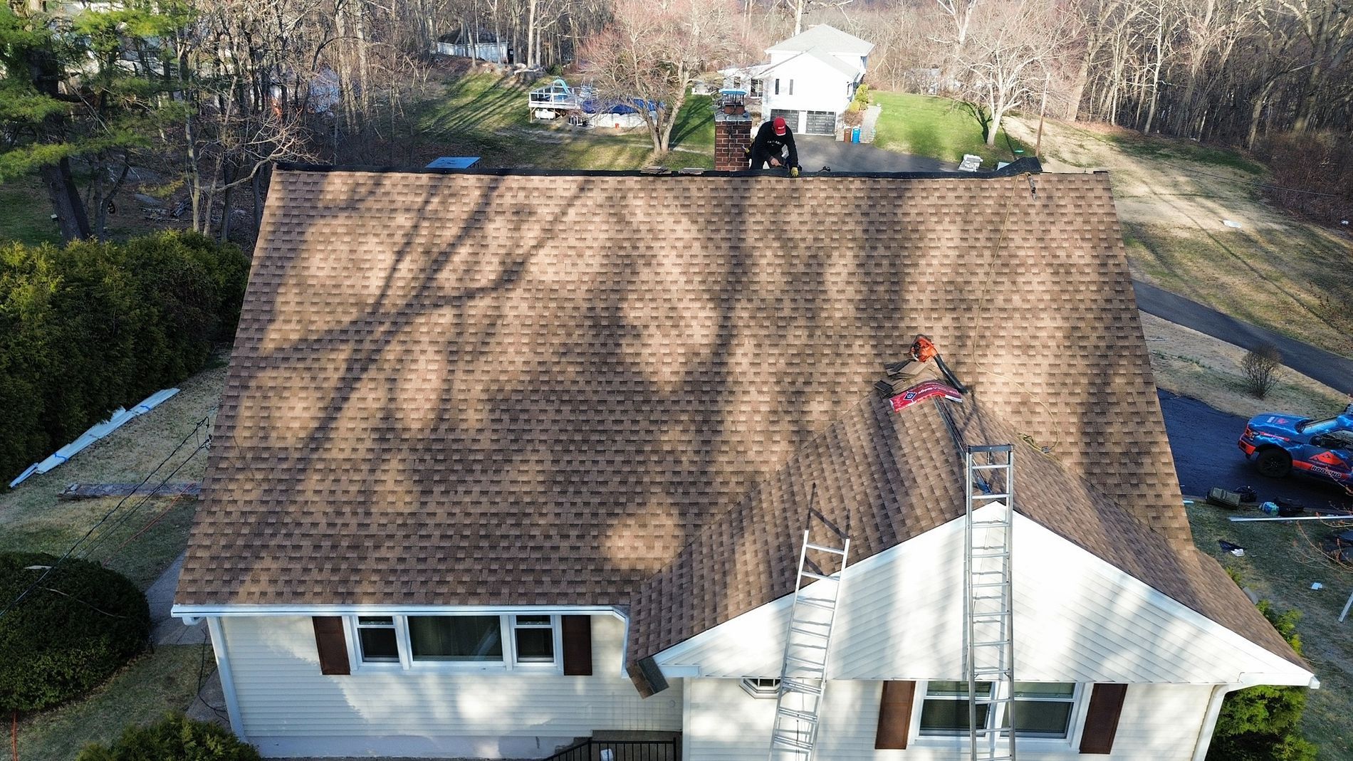 Aerial view of a house with a brown shingle roof and a ladder on the front side in a wooded yard.