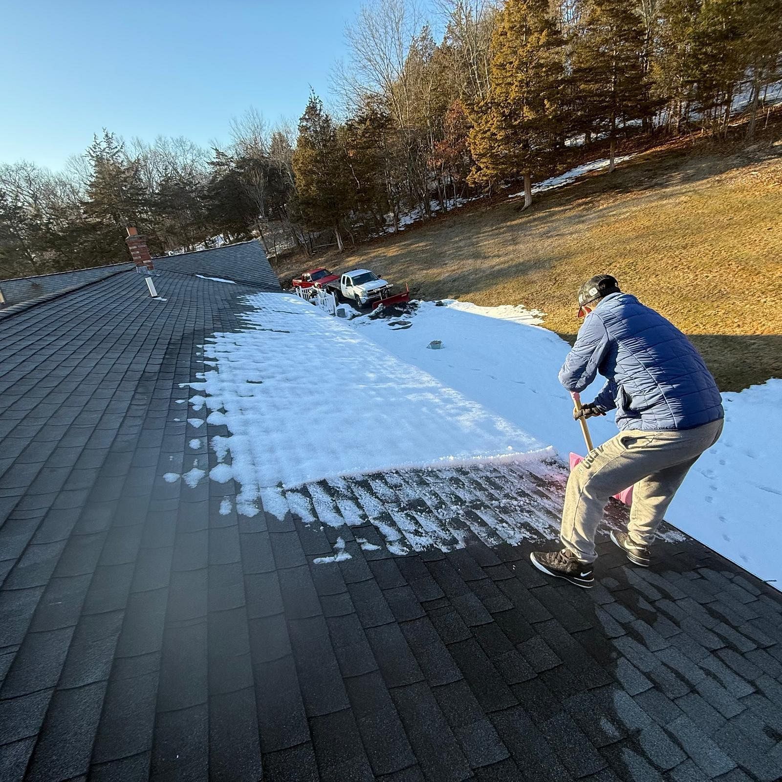 Worker spreading snow on a roof with a shovel beside a dark shingled area and pine trees