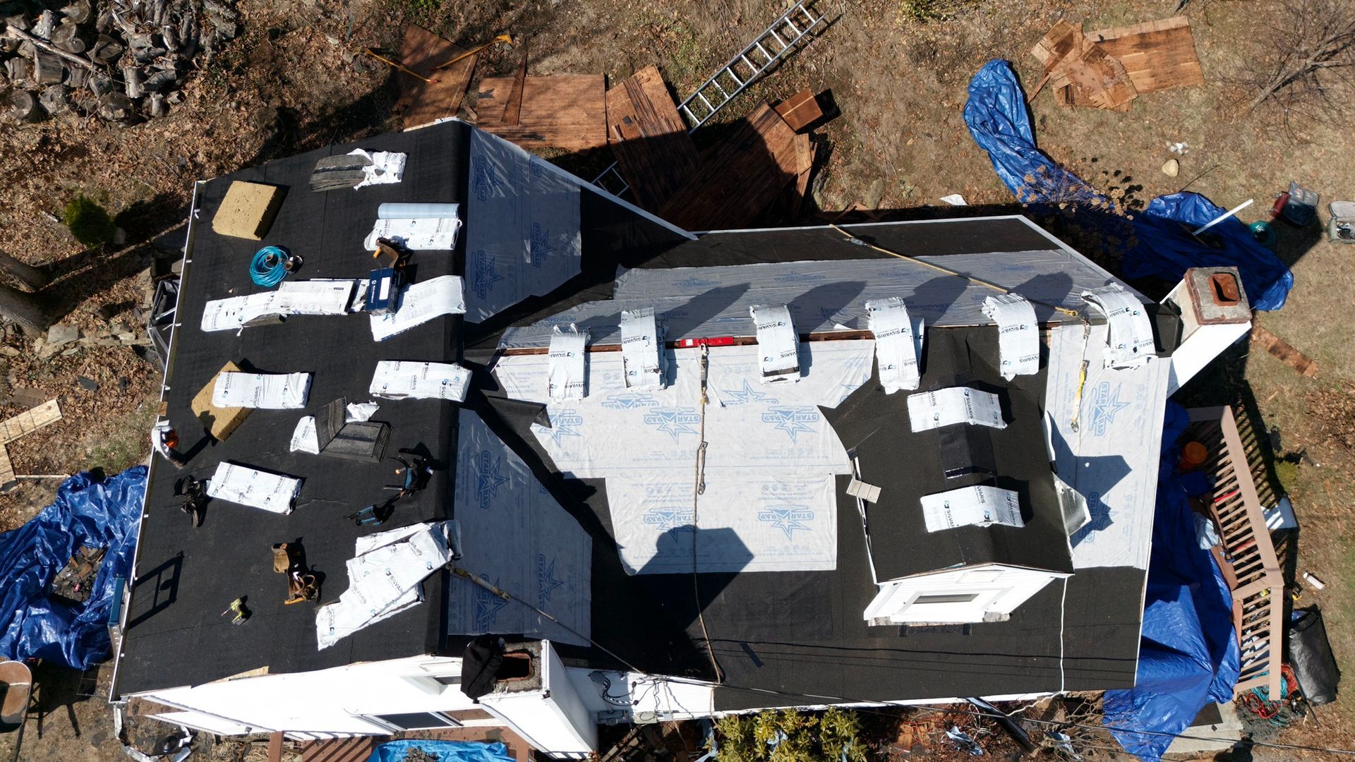Aerial view of a house roof under construction with black shingles, white underlayment, and scattered materials.