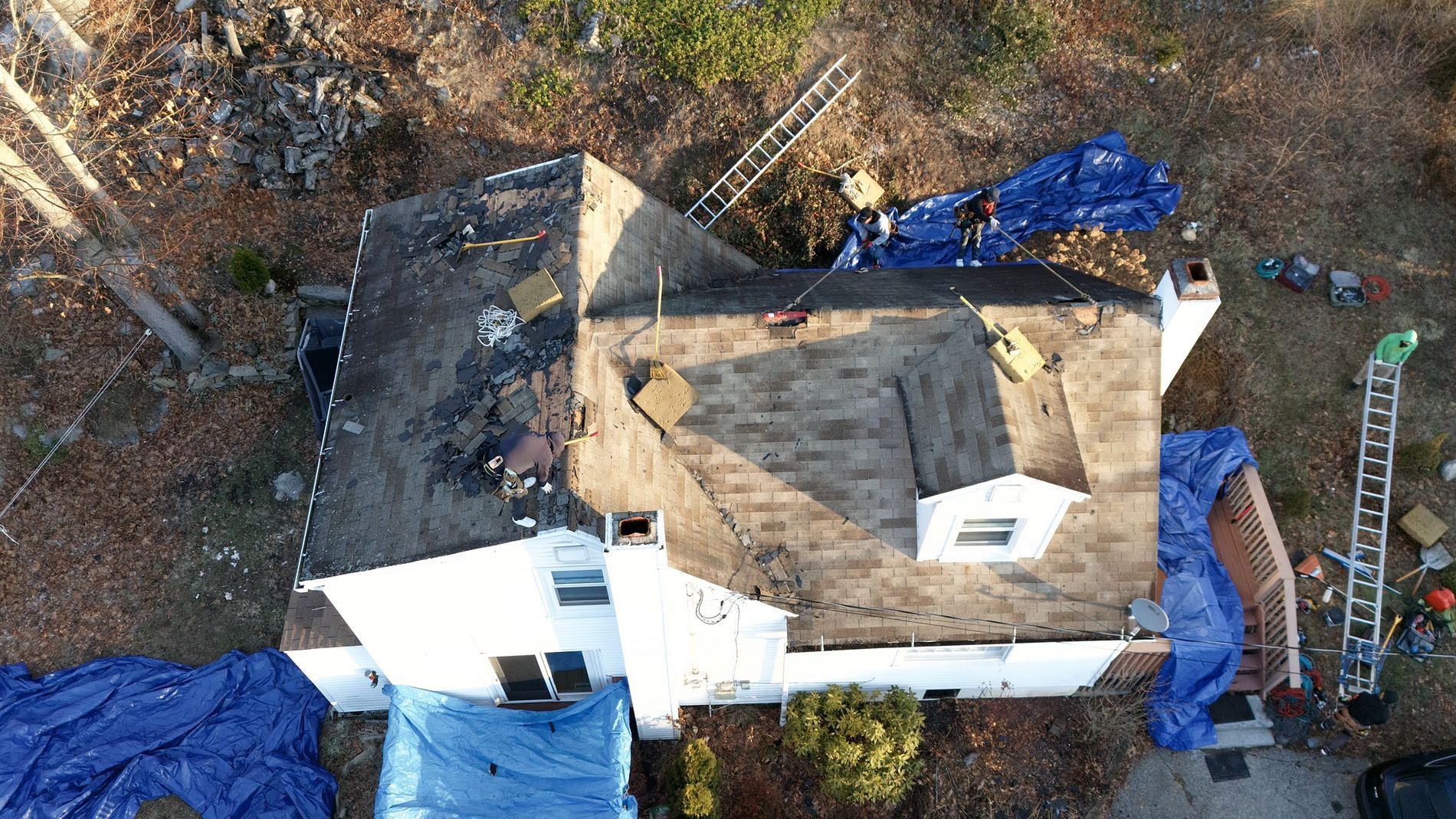 Aerial view of a house under roof repair, with blue tarps and scattered equipment around it.