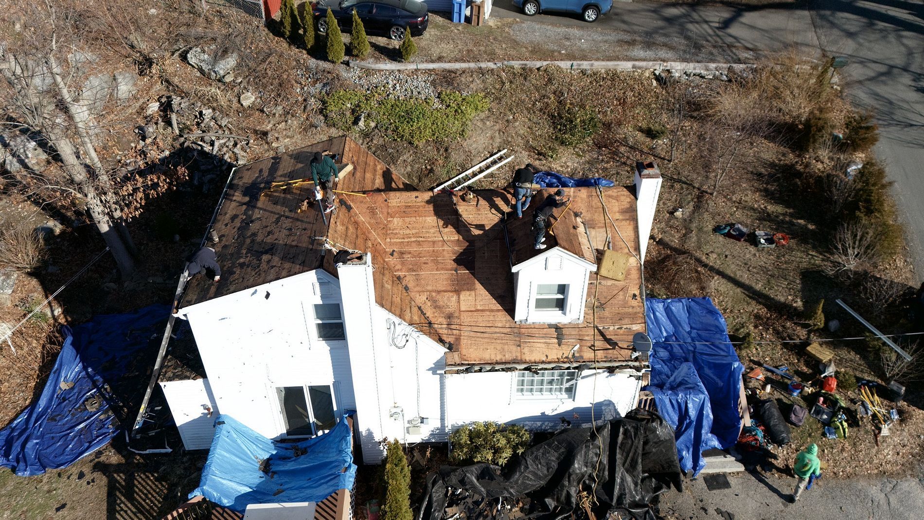 Damaged white house with a torn roof and blue tarps amid debris in a yard, aerial view