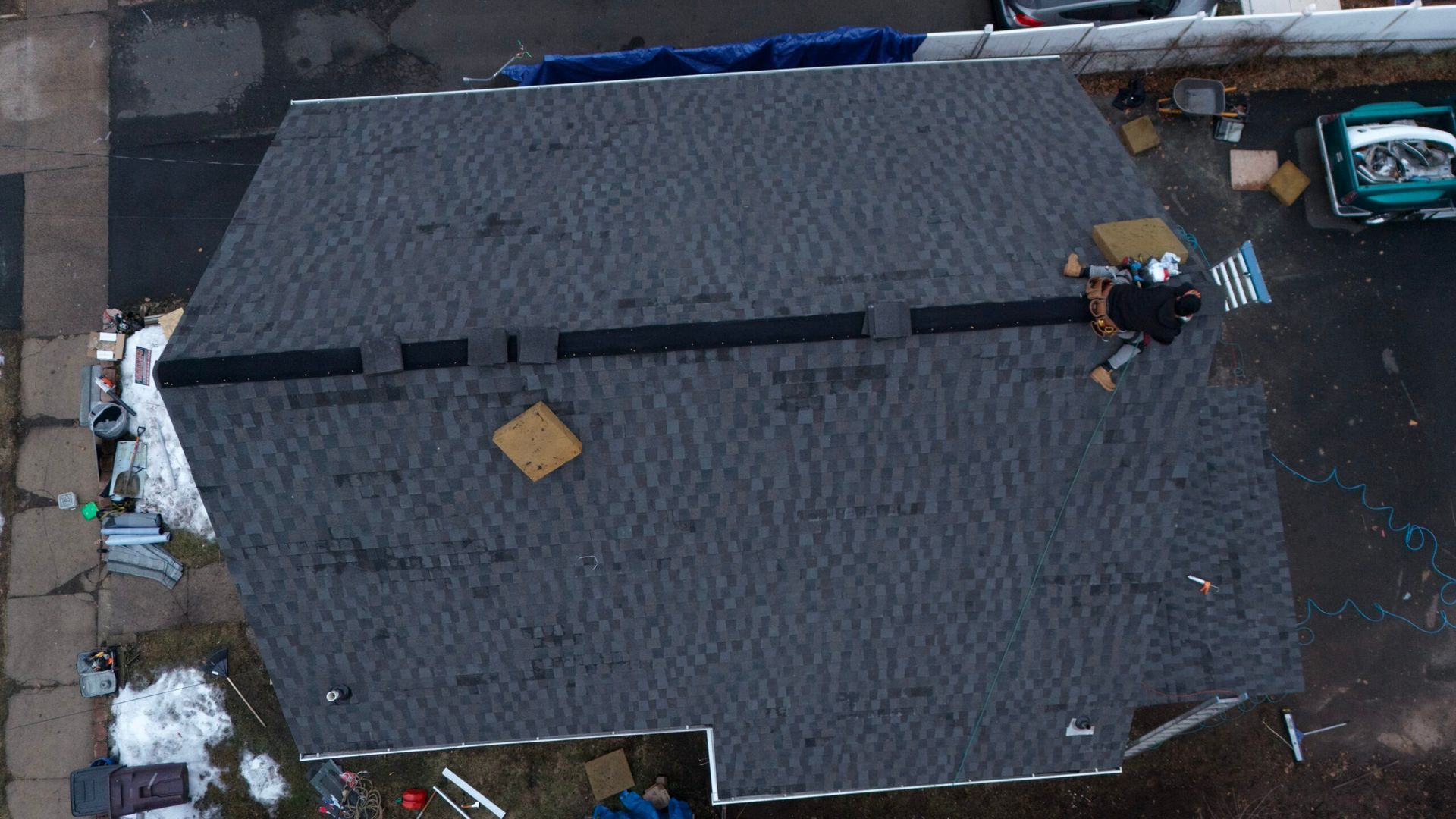 Aerial view of a dark shingled roof with workers repairing it and debris nearby.