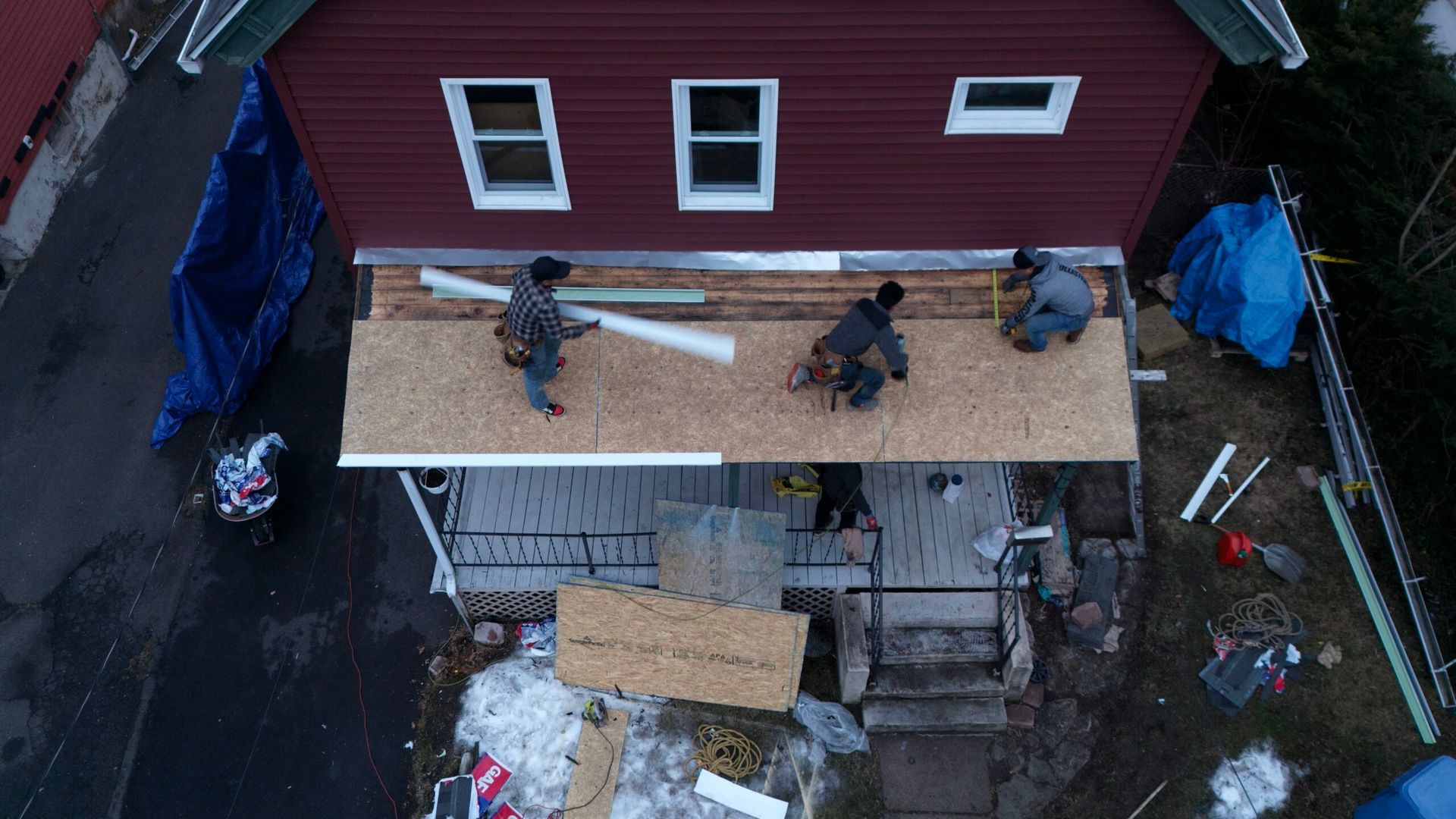 Aerial view of workers repairing a roof beside a red house with plywood and tools.