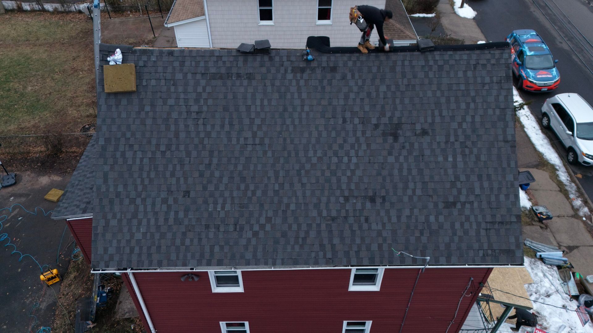 Aerial view of workers repairing a dark shingled roof on a red house, with cars parked nearby.