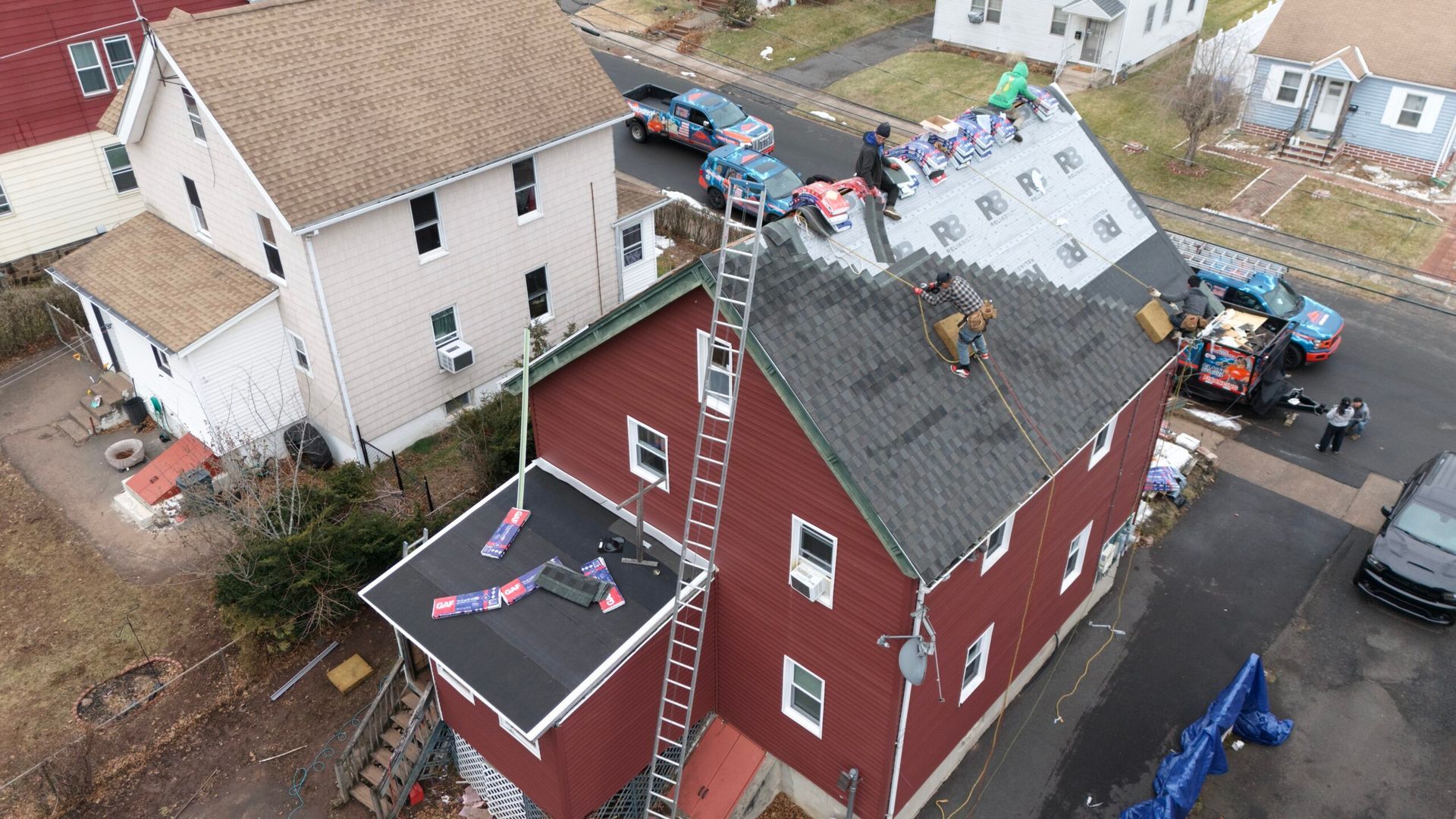 Aerial view of roofers repairing storm damage on a red house beside a white house and work trucks.