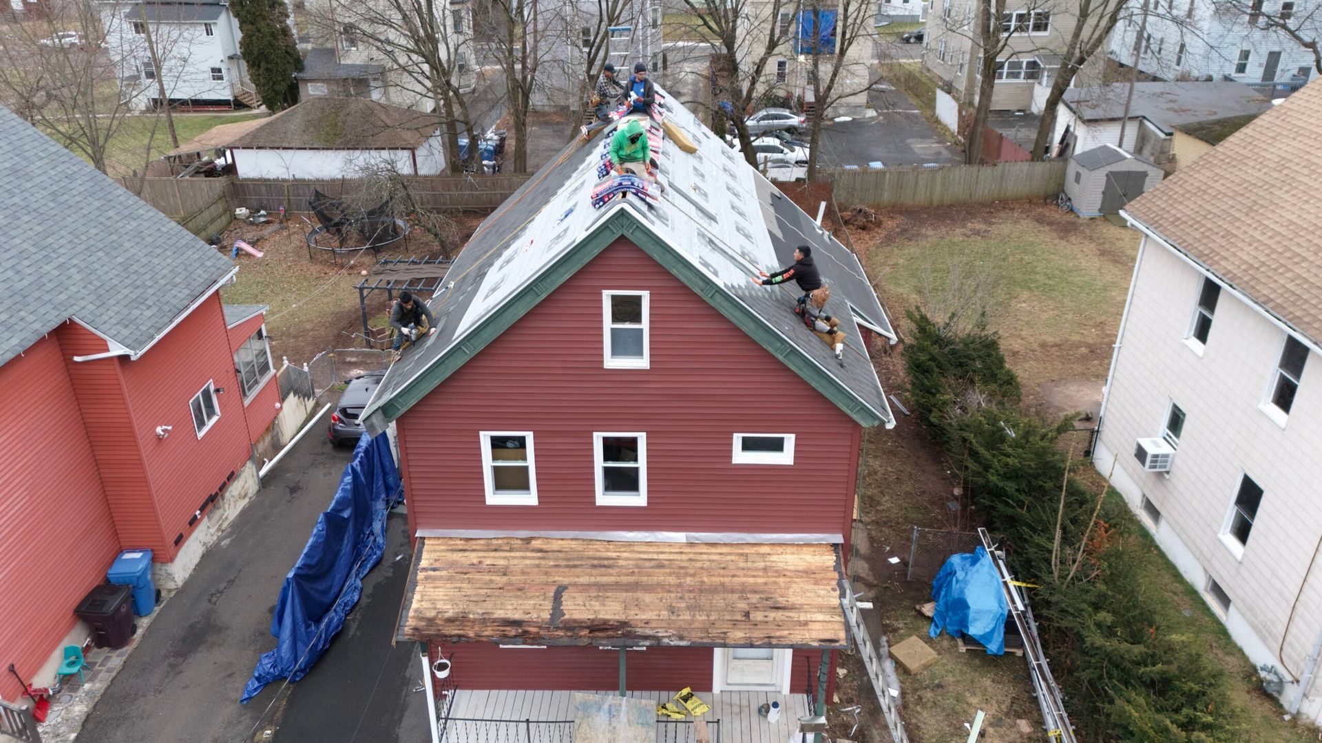 Aerial view of a red house with a partially repaired roof, flanked by neighboring homes and backyard tarps.