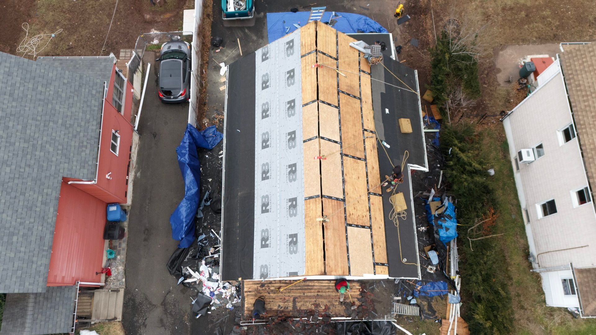 Aerial view of a roof under construction, covered with tan plywood and blue tarps between neighboring houses.
