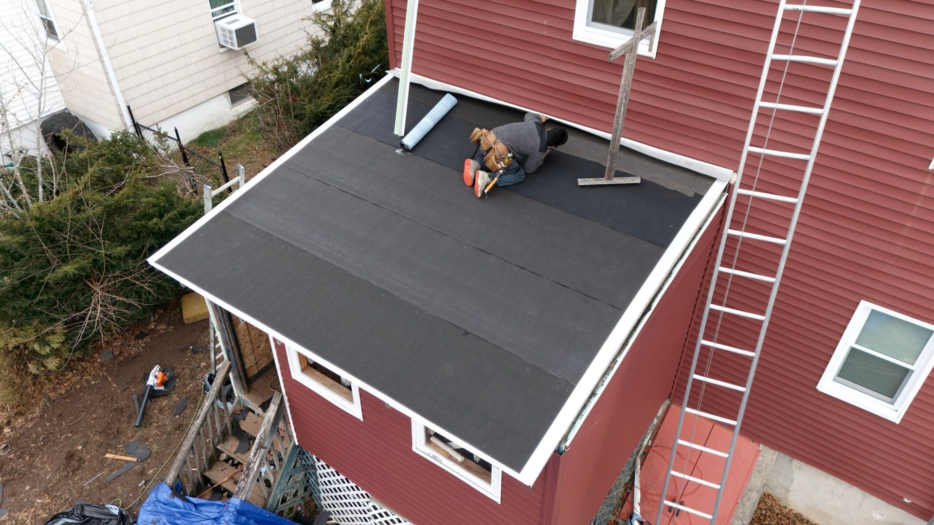 Worker installing a black roof on a red house beside a ladder