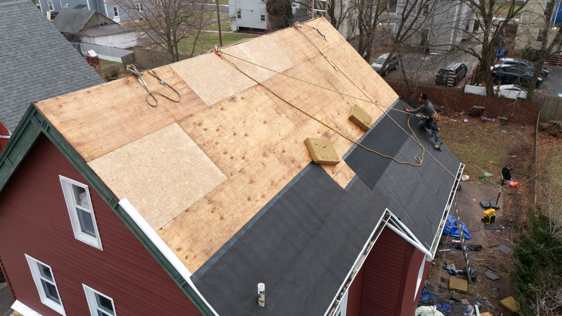 Aerial view of a red house with a partially repaired roof covered in plywood and black shingles.