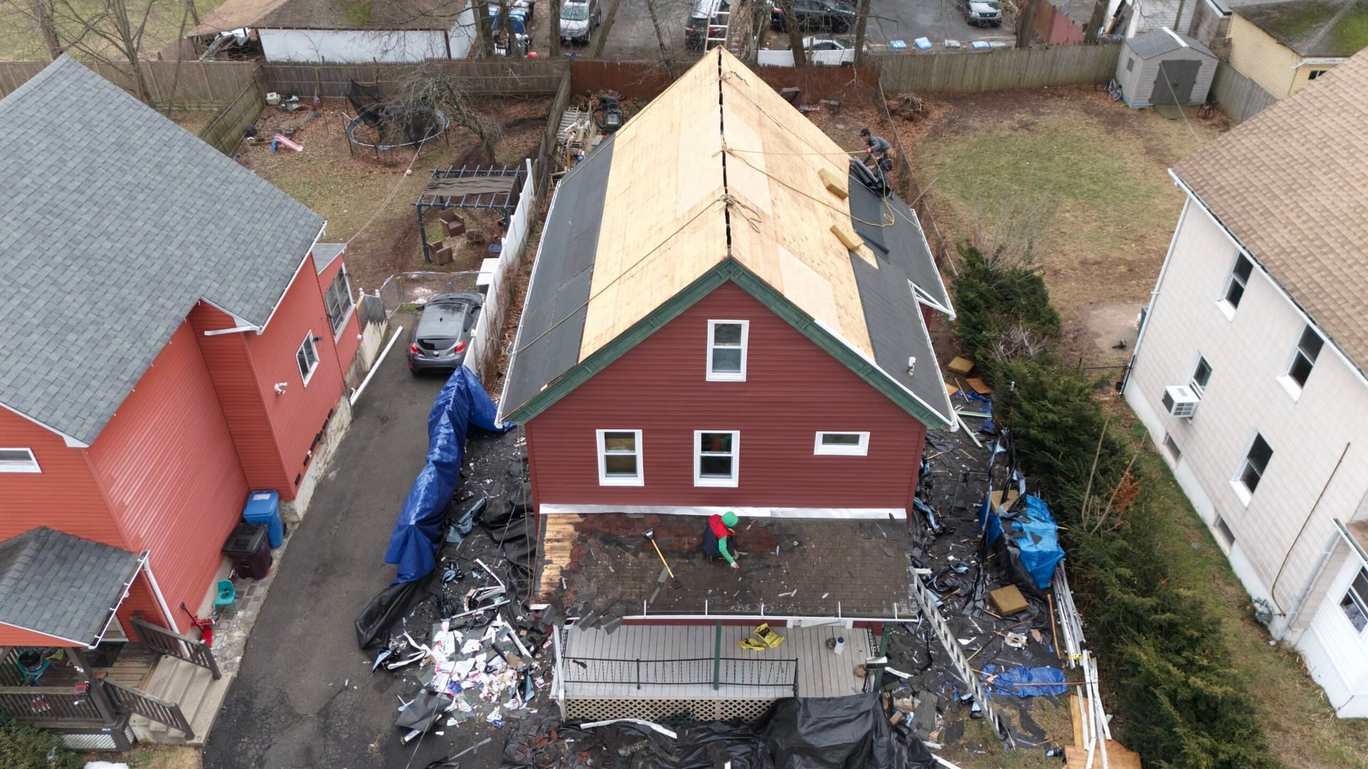 Aerial view of a red house with a new roof under construction, surrounded by debris and nearby homes