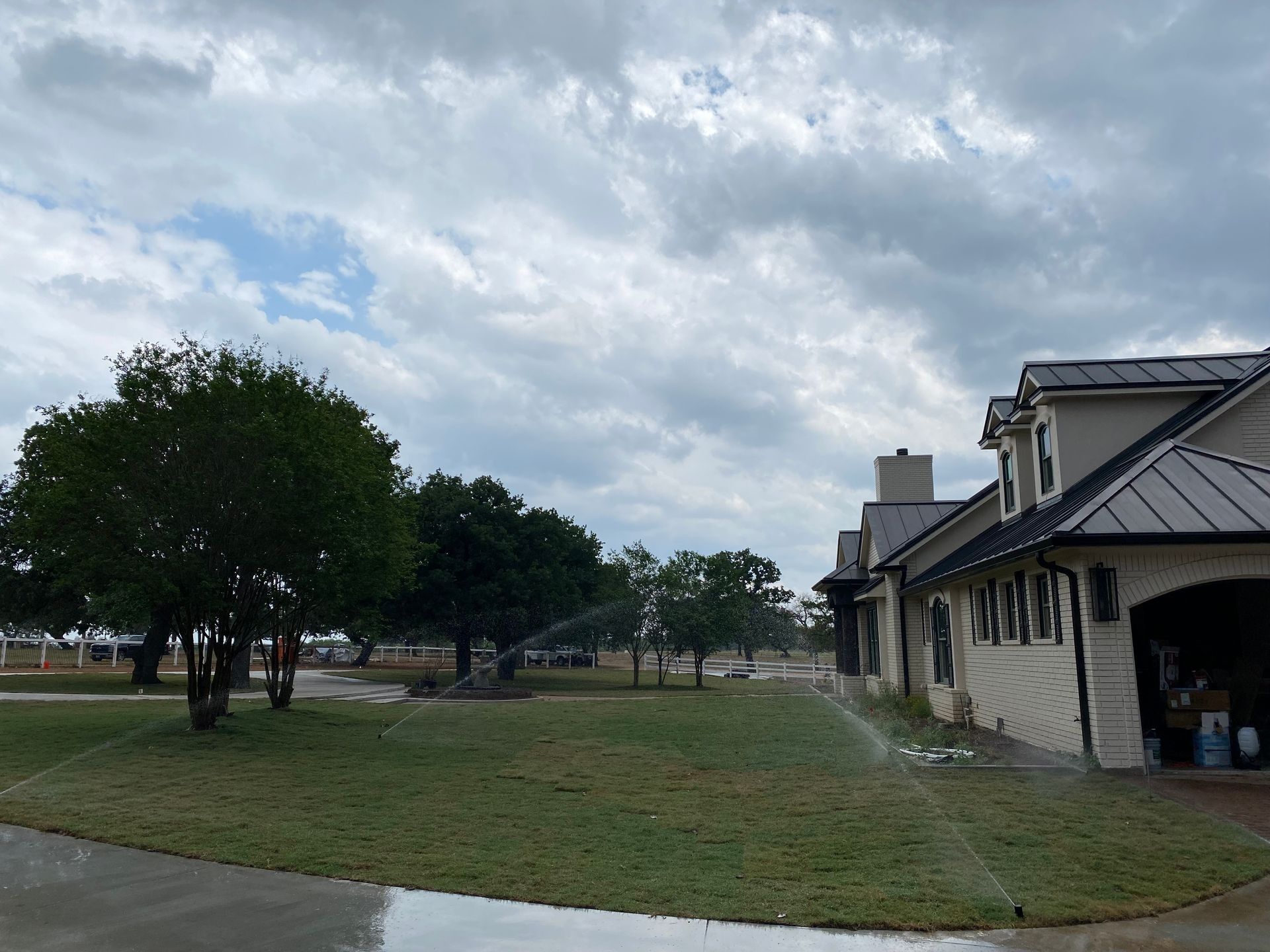 Sprinklers watering green lawn near a two-story beige house under a cloudy sky.