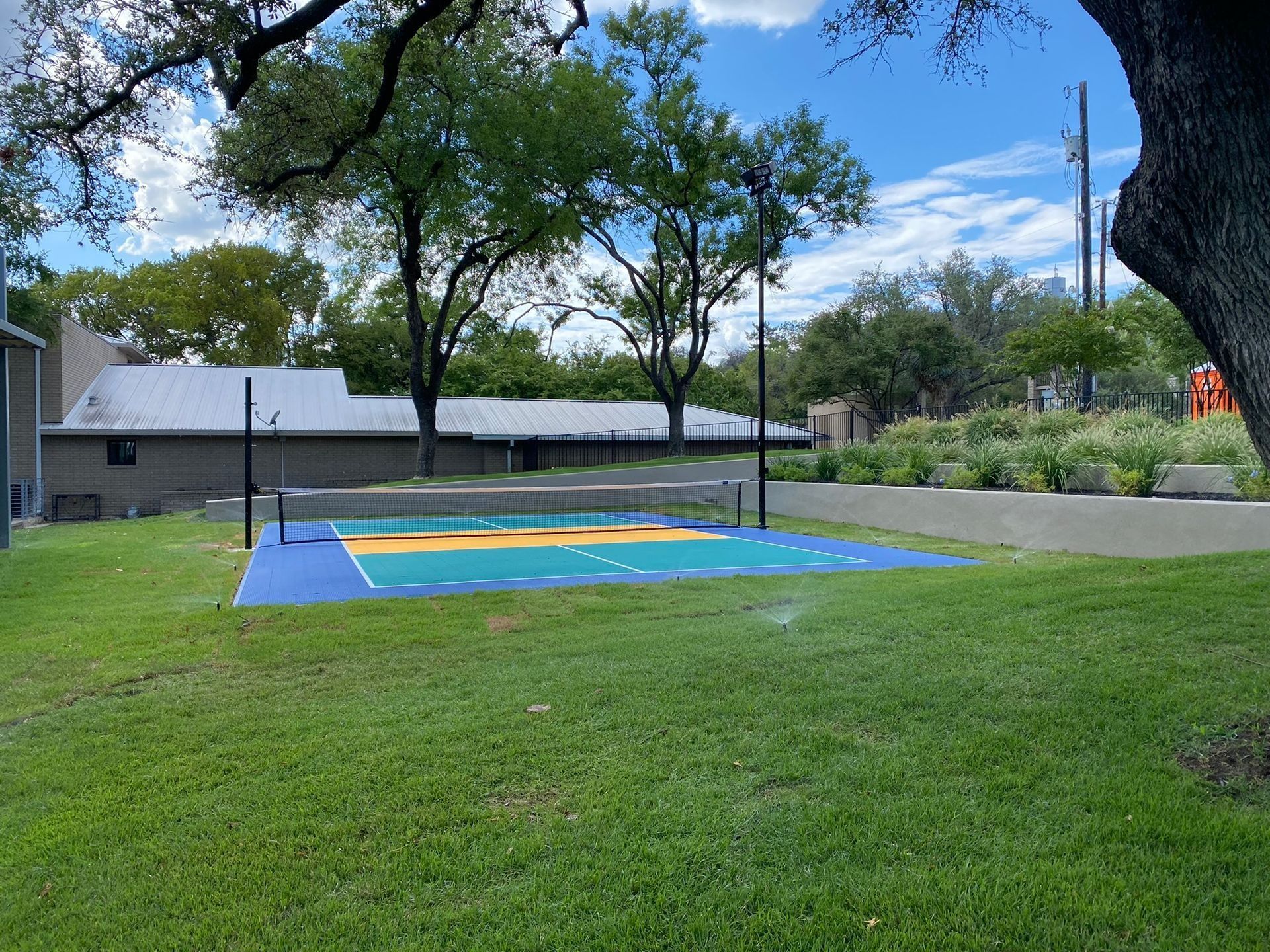 Volleyball court with net, colorful painted surface, green grass, trees, and a building in the background.