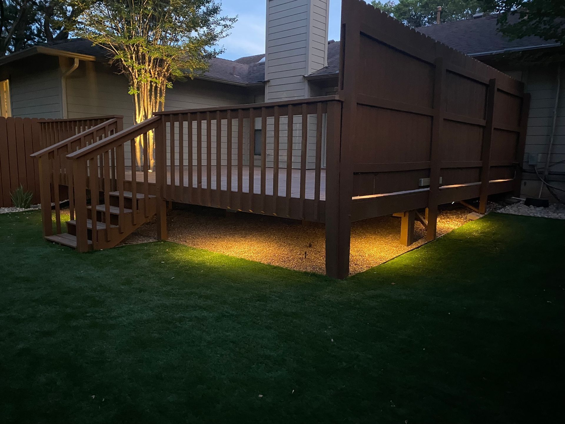 Wooden deck with stair, lit by warm lights, on green grass, in backyard.
