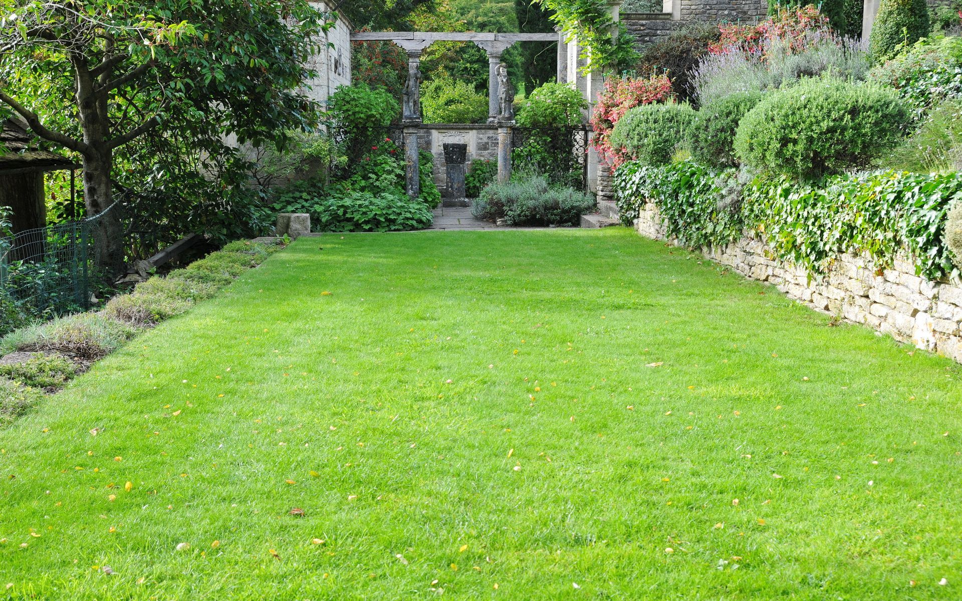 Lush green lawn in a garden, leading to a stone structure with archways and lush foliage.