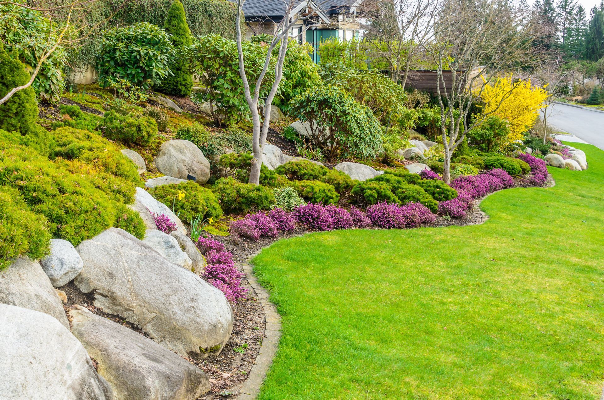Landscaped garden bed with large rocks, colorful plants, and green lawn.