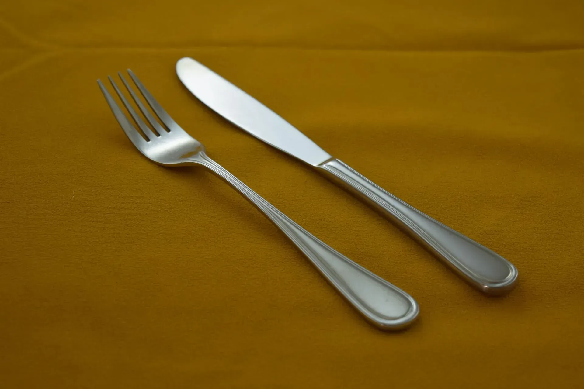 A silver fork and a silver knife placed diagonally on a plain yellow tablecloth.