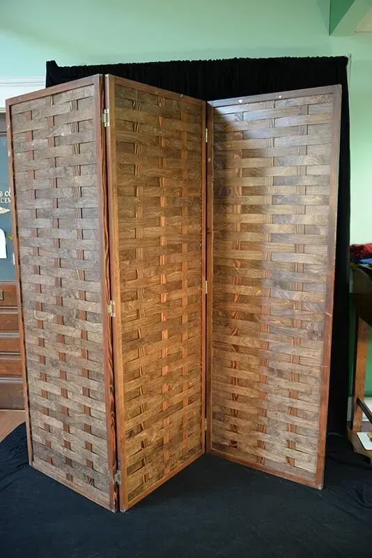 A three-panel folding room divider with a brown, woven wooden lattice pattern set against a black backdrop.