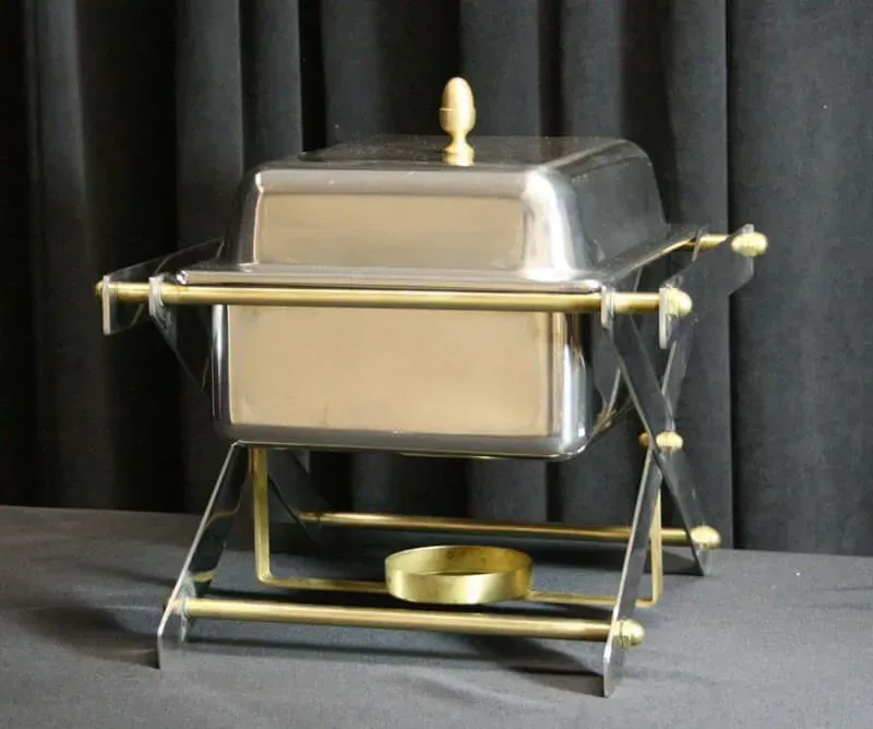Stainless steel chafing dish on a stand with a gold handle and cover knob, set against a black backdrop