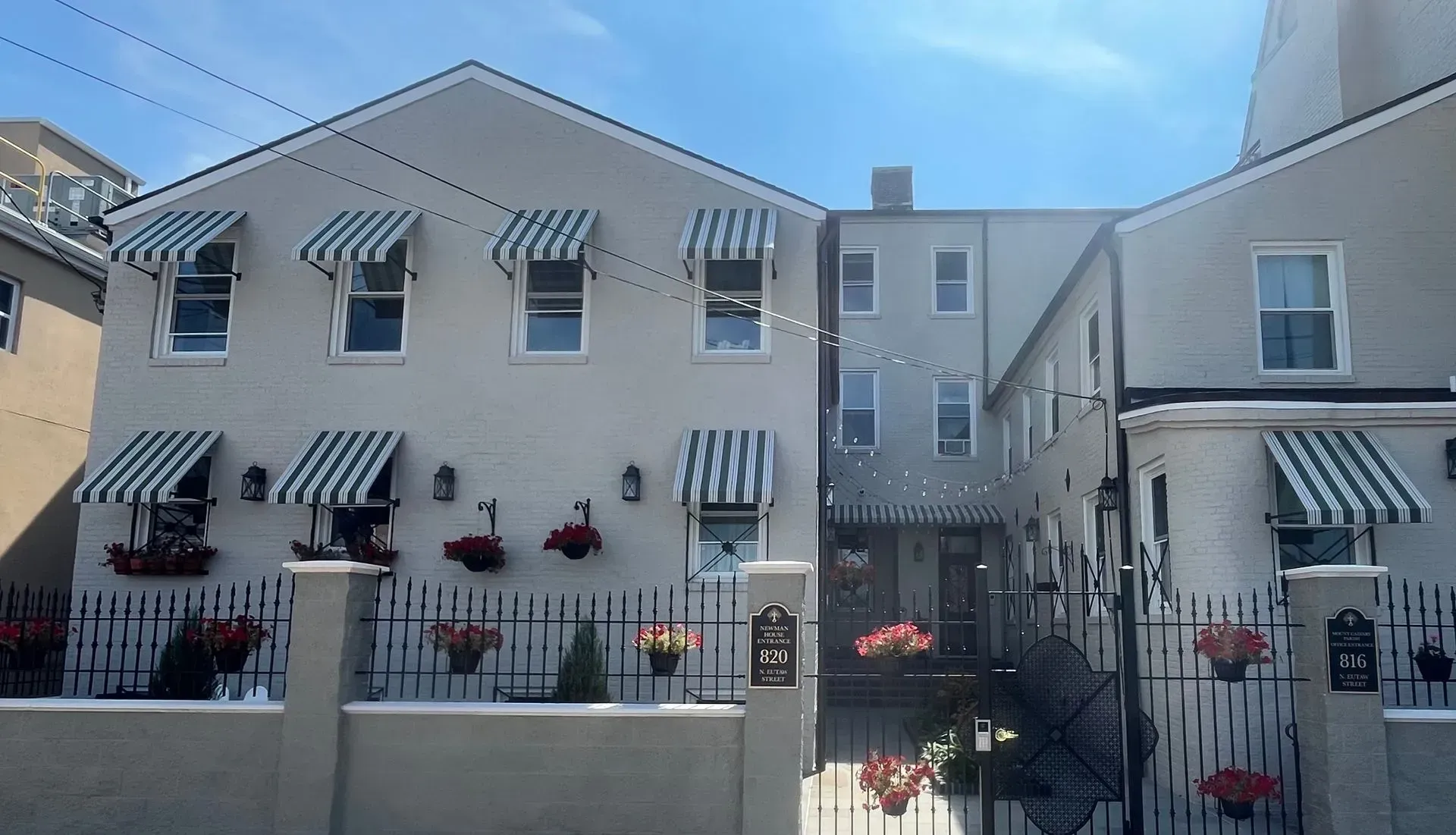 A two-story light gray building with green and white striped awnings over windows, behind a gated courtyard with planters.