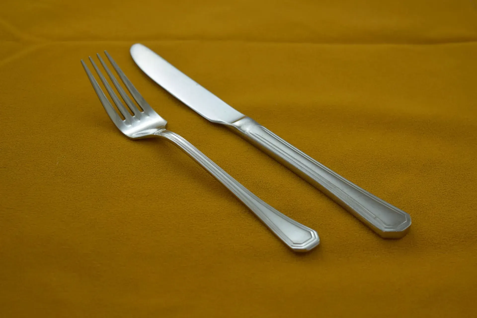 A silver fork and dinner knife laid side-by-side on a mustard-yellow fabric background.