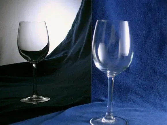 A clear wine glass stands on a blue cloth, with its reflection appearing on a dark surface to the left.