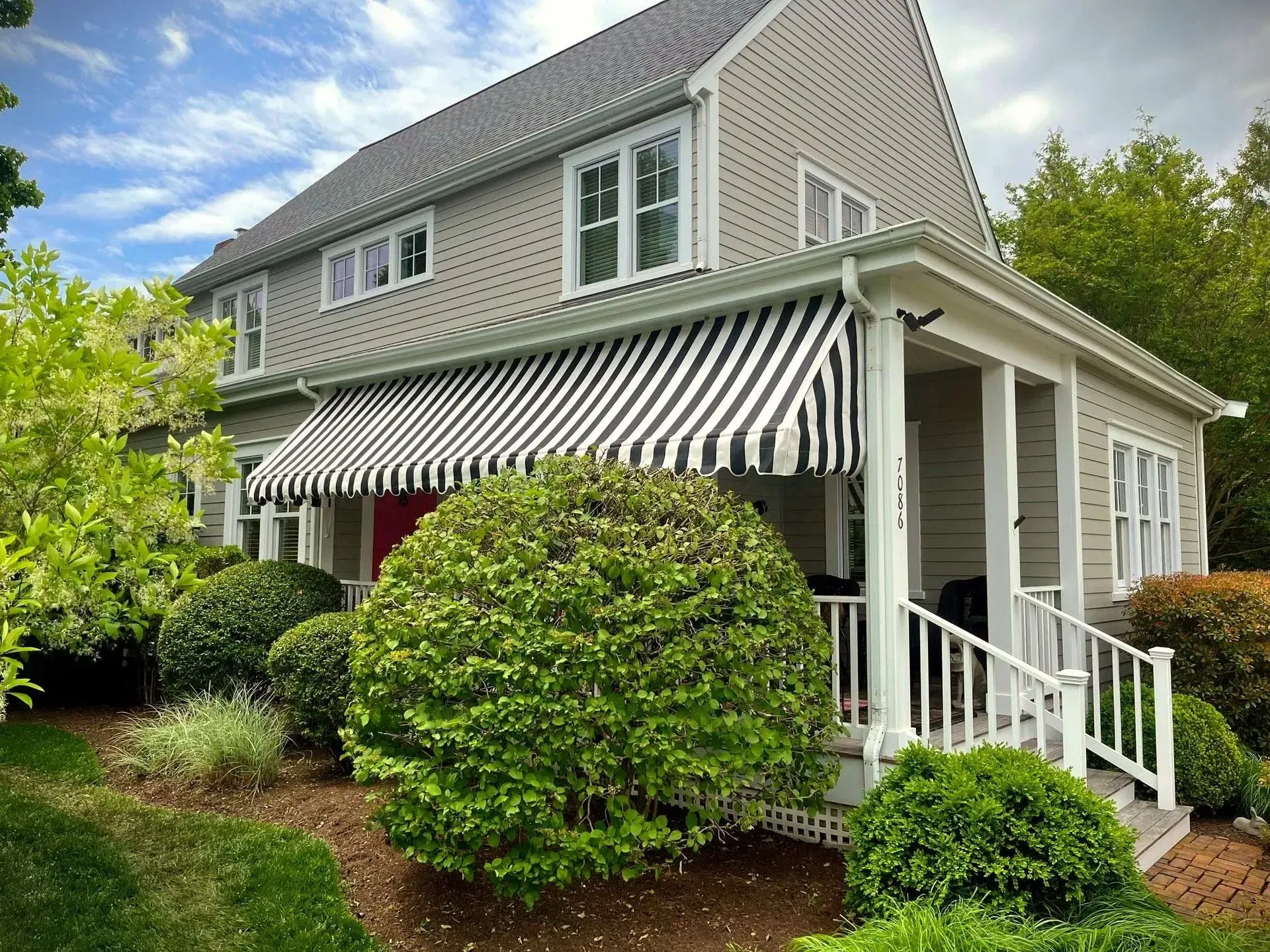 A light gray two-story house with a white porch, black-and-white striped awning, and lush green landscaping.