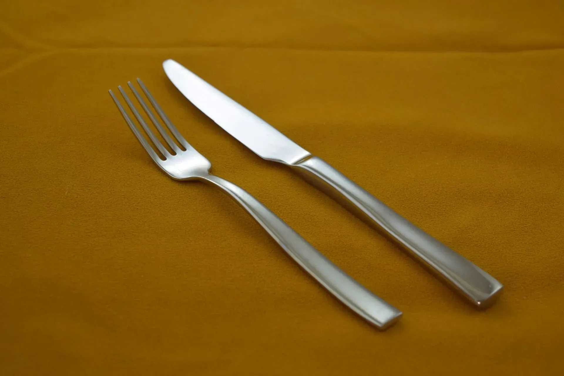 A silver fork and a silver dinner knife laid side-by-side on a plain, dark gold-colored tablecloth.