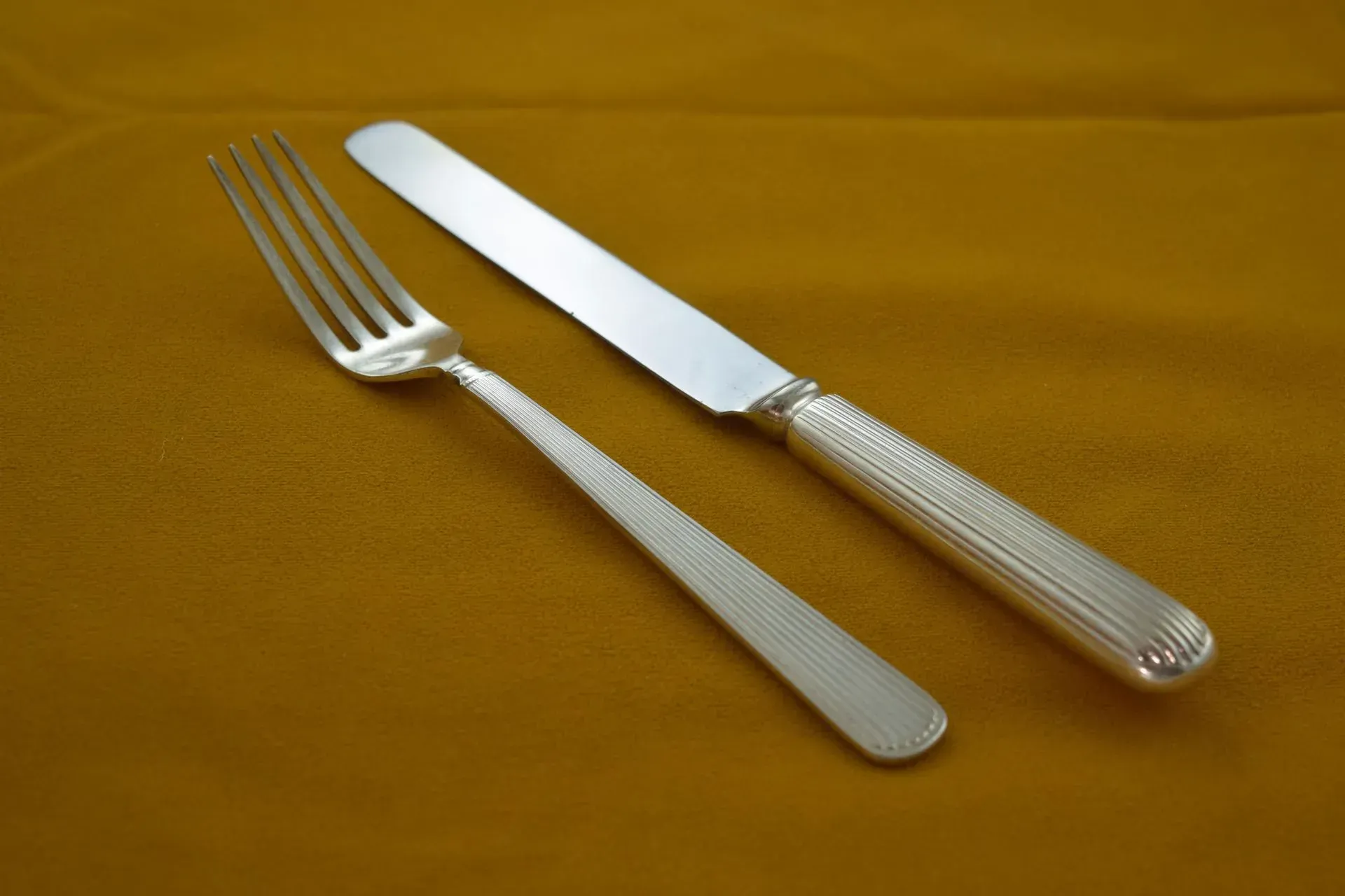 A silver fork and dinner knife with a ribbed handle design, positioned diagonally on a plain, ochre-colored background.