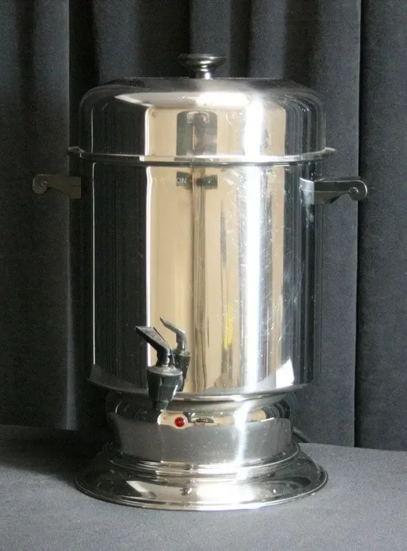 Shiny silver stainless steel beverage dispenser with spigot on a gray surface against a dark curtain.