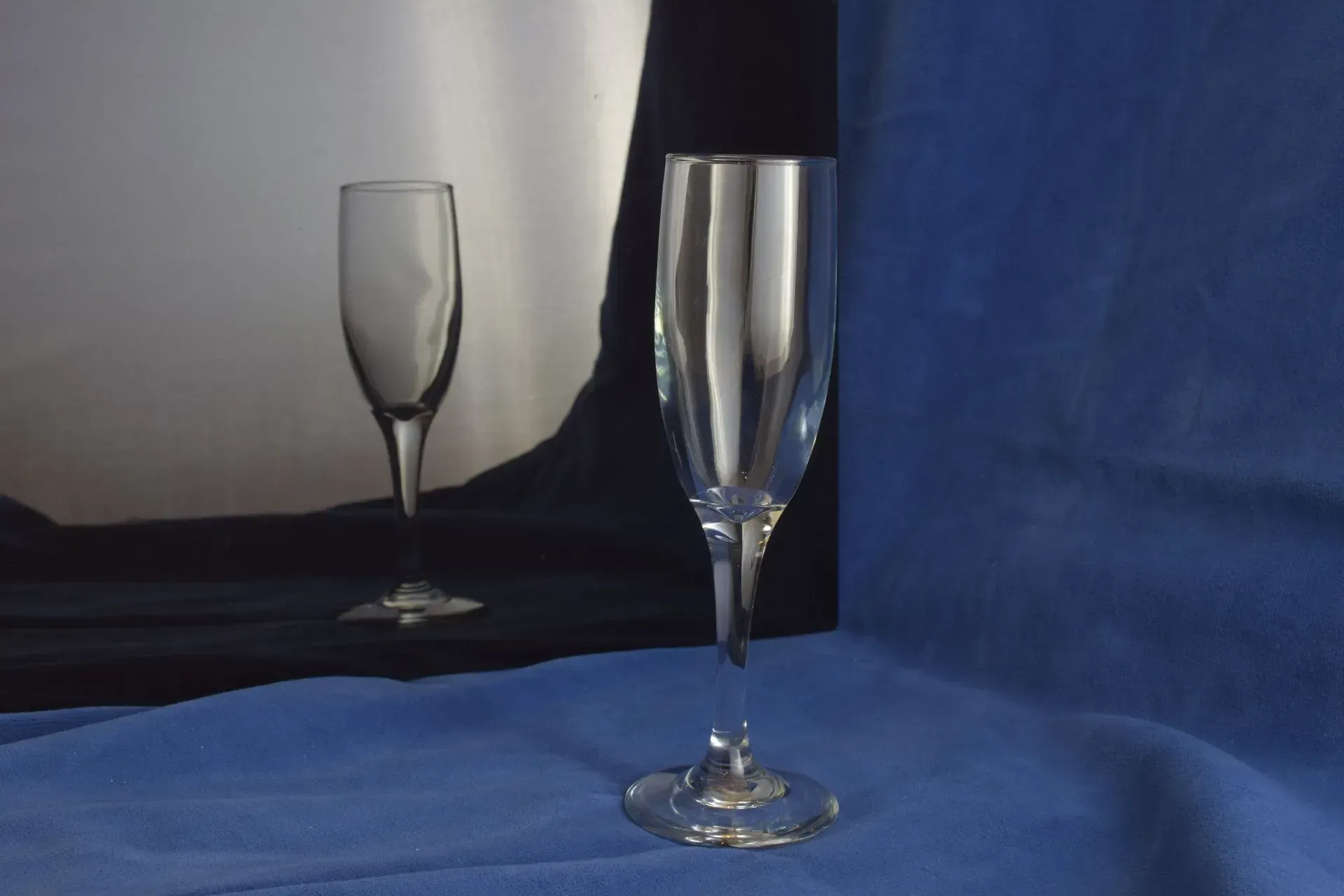 A clear glass champagne flute sits on a blue cloth, with its reflection visible in a mirror behind it.