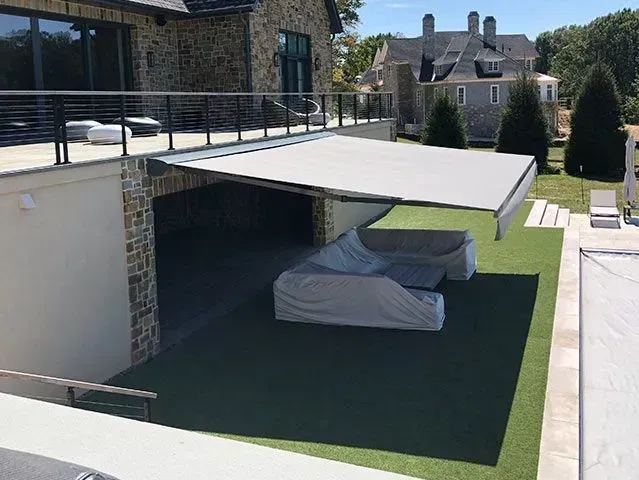 An outdoor patio area with a retractable beige awning covering a grey sofa set on a green artificial turf lawn.