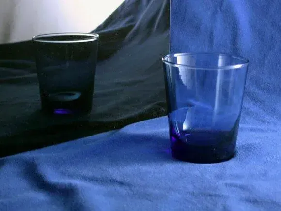 A blue glass rests on a blue cloth, with a mirror reflecting the glass onto a black surface.