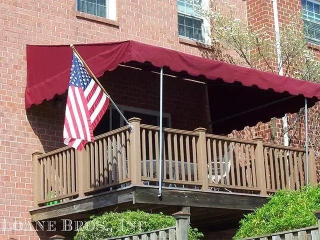 A maroon canvas awning over a wooden balcony on a brick building, with an American flag attached to a support pole.