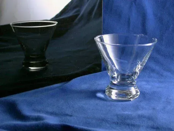 A clear glass cocktail bowl sits on a blue fabric surface in front of a mirror reflecting a black background.