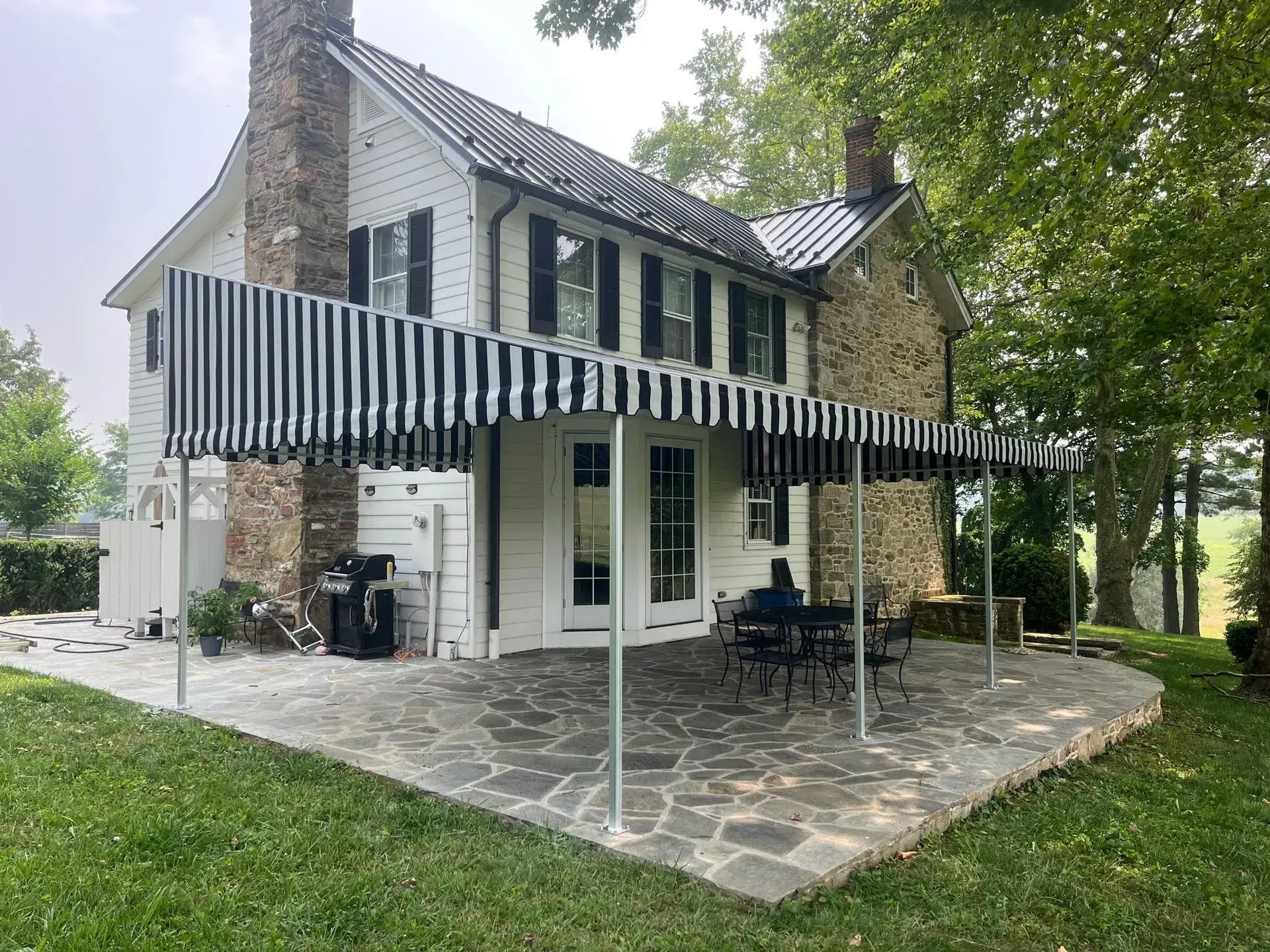 A white house with a stone chimney and a black-and-white striped awning over a stone patio with outdoor seating.