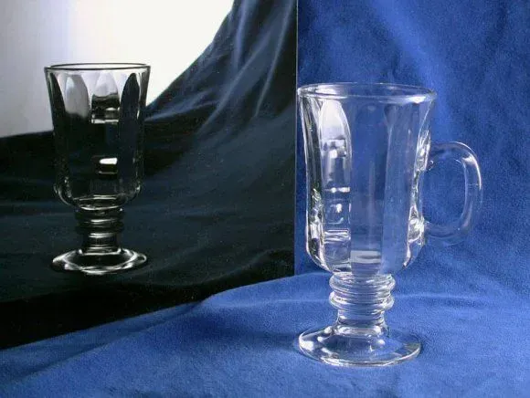 A clear glass Irish coffee mug with a handled, footed base resting on a blue cloth, reflected in a mirror.