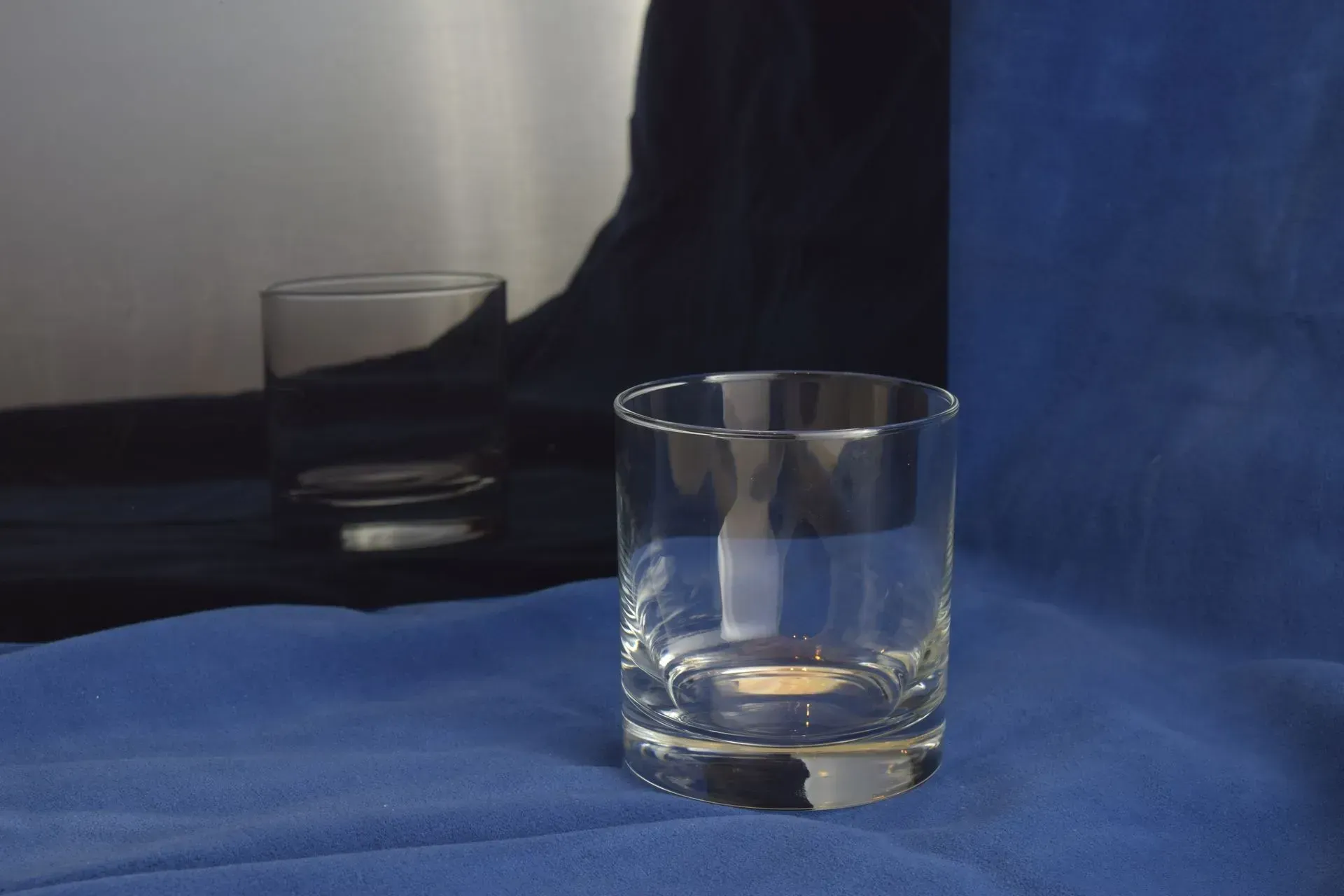 Two clear glass tumblers sit on a blue fabric surface, with one in focus in the foreground and one blurred behind it.