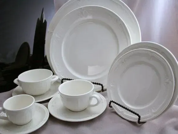 A set of white ceramic dinnerware, including plates, saucers, and cups, arranged on a neutral fabric surface.