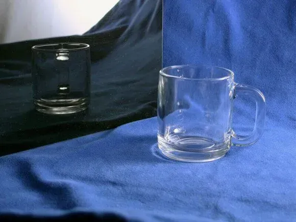 A clear glass mug sits on a blue fabric surface, reflected in a mirror placed on a dark background.