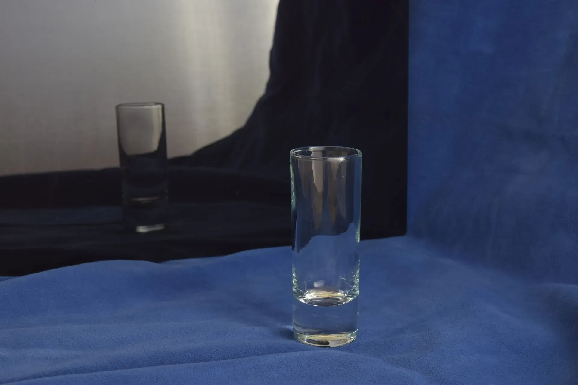 A clear shot glass stands on a textured blue surface with a mirror reflection of the glass visible in the background.