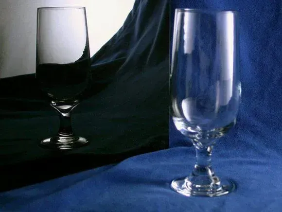 A clear glass stands on a blue cloth, showing a distinct dark reflection of itself on a smooth black surface.
