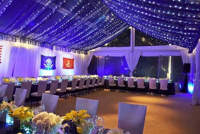 A banquet tent decorated with purple string lights, white draped fabric, and two flags, set with long dining tables.